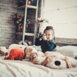 Smiling child with a camera surrounded by stuffed toys in a cozy bedroom setting.